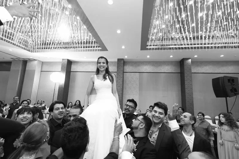Black and white photograph of a bride in a white gown lifted on a chair by cheering guests at the reception