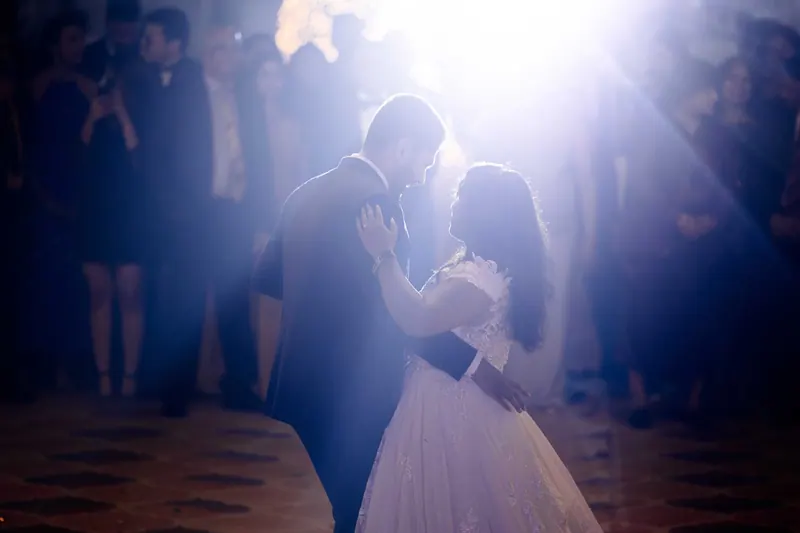 Couple's first dance in a beam of warm stage light, guests watching in silhouette