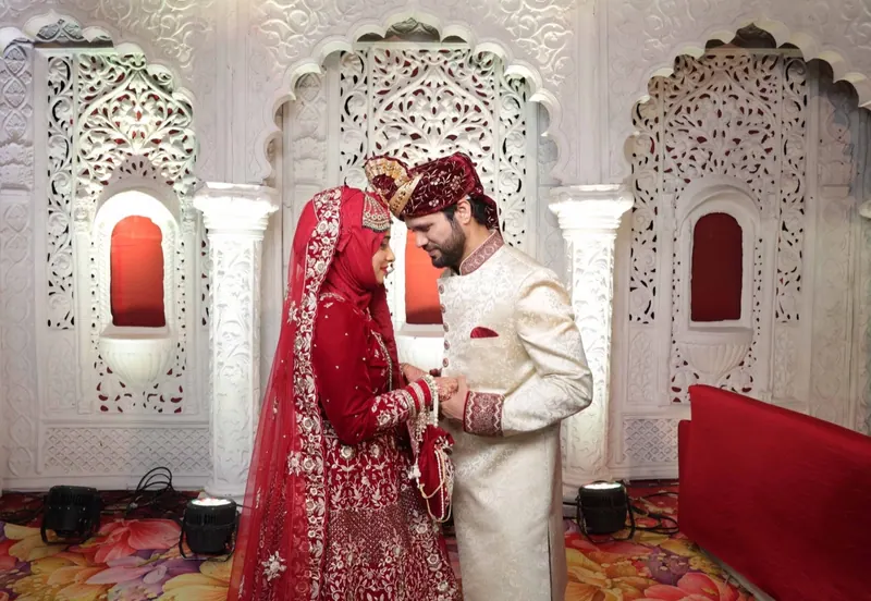 Bride in a red dupatta and groom in an ivory sherwani hold hands beneath carved white arches